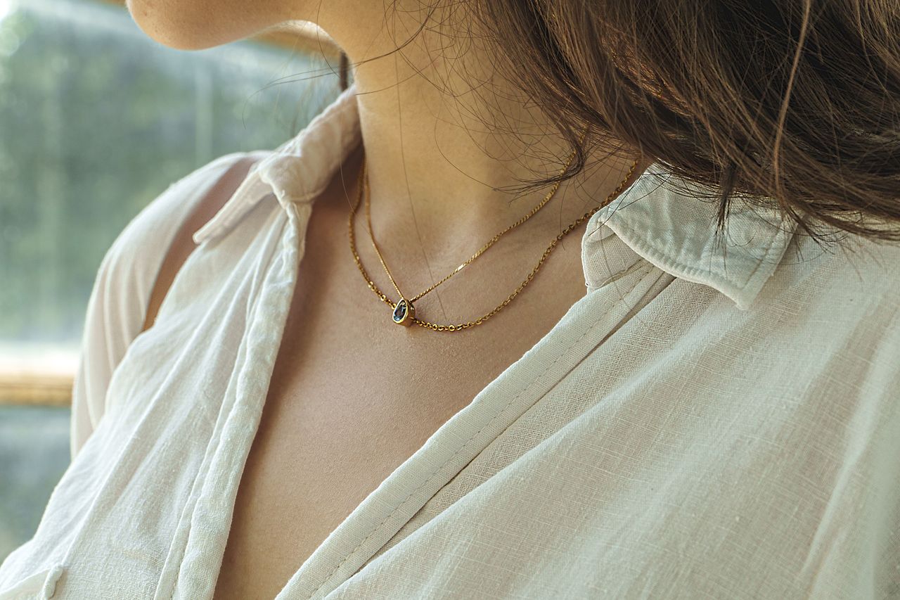 A close-up portrait of a woman in an elegant white blouse wearing a delicate gold necklace with a pendant.